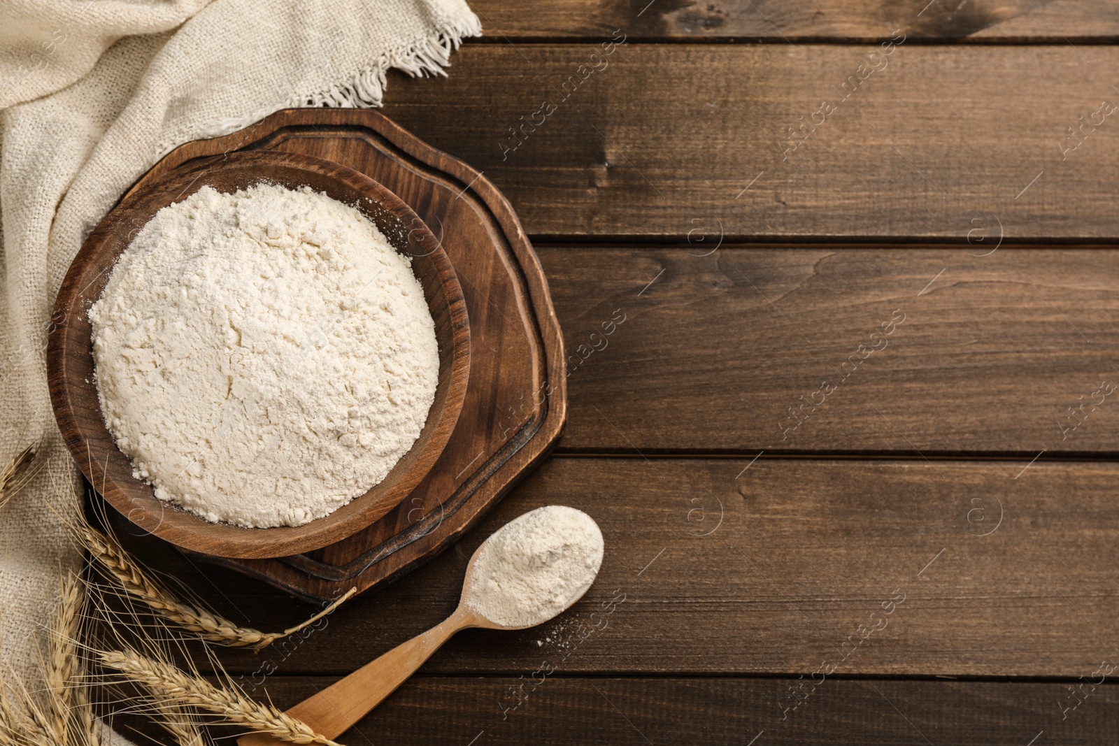 Photo of Bowl with wheat flour, spoon and spikelets on wooden table, flat lay. Space for text