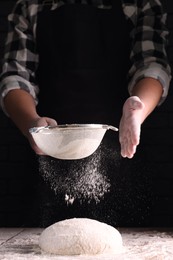 Photo of Man sprinkling flour over dough at wooden table on dark background, closeup
