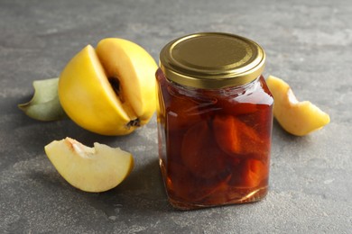 Photo of Tasty homemade quince jam in jar and fruits on grey textured table, closeup