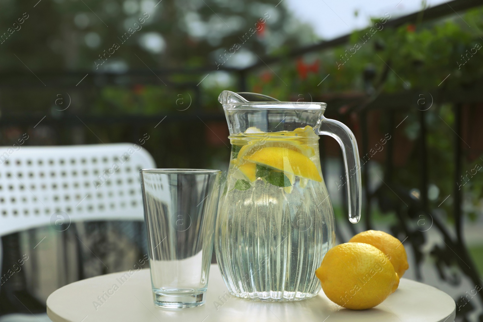 Photo of Jug with refreshing lemon water, glass and citrus fruits on light table outdoors