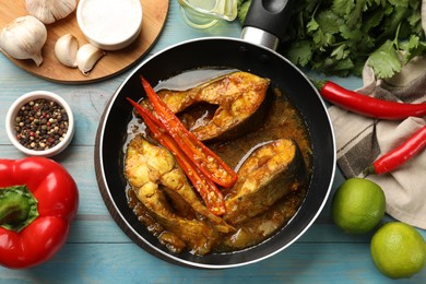 Photo of Tasty fish curry in frying pan and ingredients on light blue wooden table, flat lay. Indian cuisine