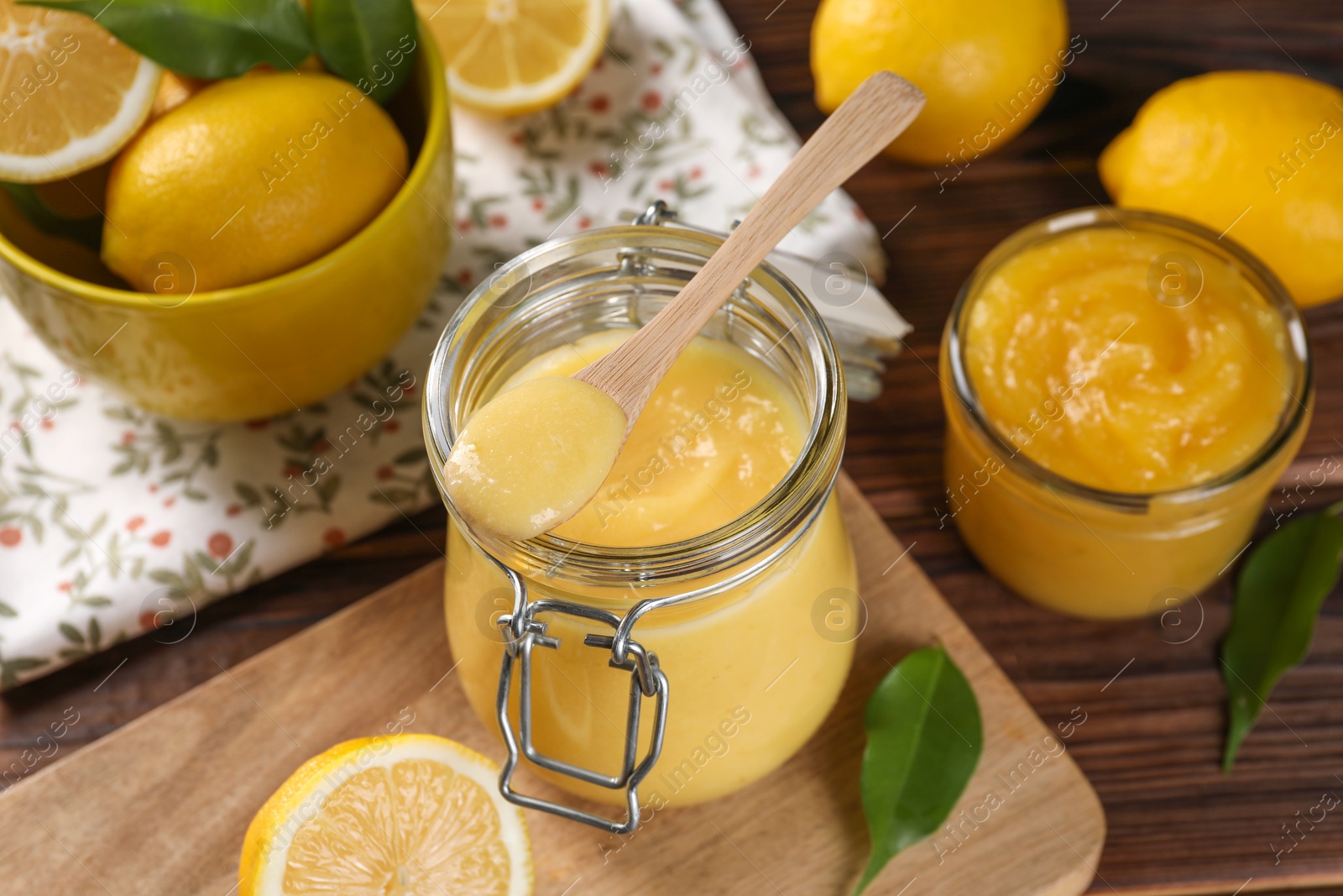 Photo of Delicious lemon curd in glass jars, spoon, fresh citrus fruits and green leaves on wooden table