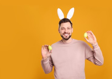 Photo of Happy man in bunny ears headband holding painted Easter eggs on orange background. Space for text