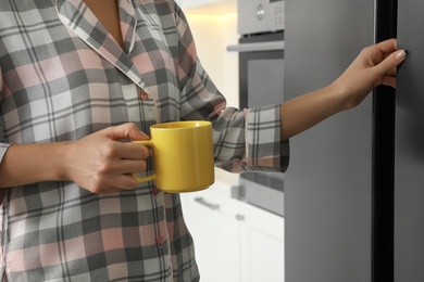 Woman with cup opening refrigerator at home, closeup