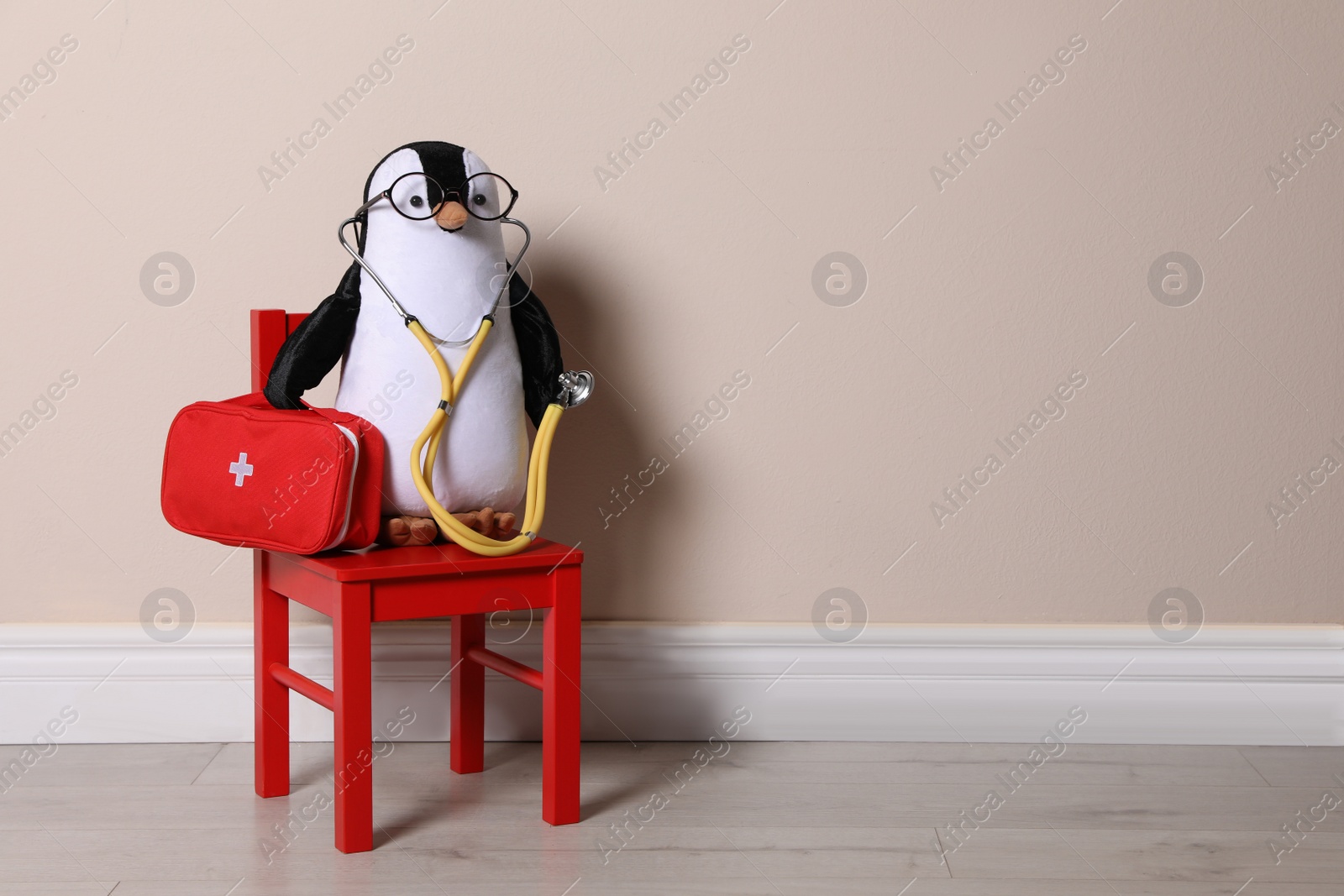 Photo of Toy penguin with eyeglasses, stethoscope and first aid bag near beige wall, space for text. Pediatrician practice