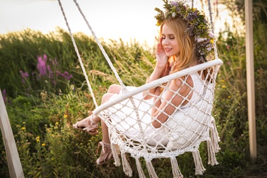 Young woman wearing wreath made of beautiful flowers on swing chair outdoors