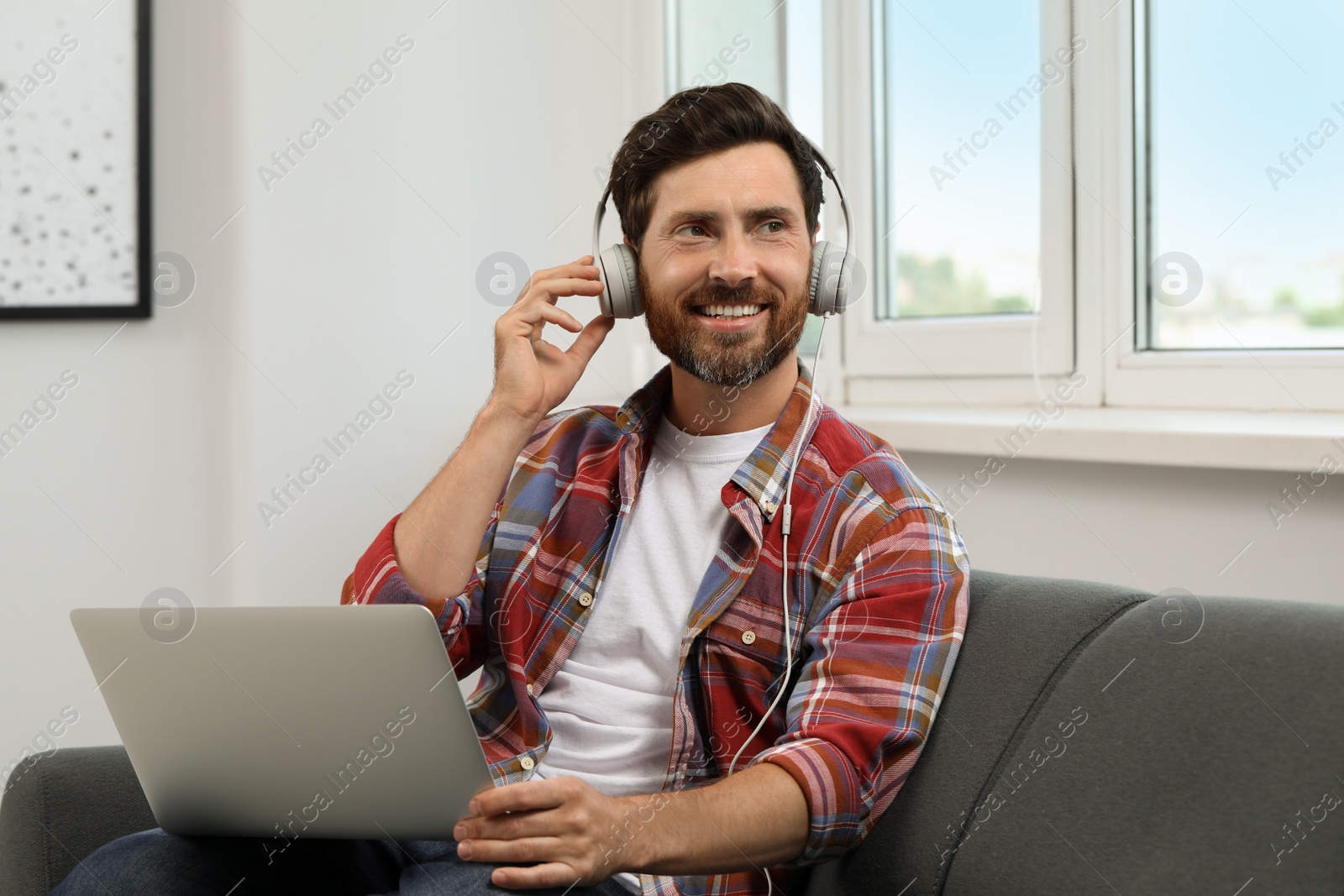 Photo of Smiling bearded man with headphones and laptop on sofa indoors