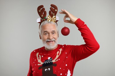 Senior man in Christmas sweater and reindeer headband holding festive bauble on grey background