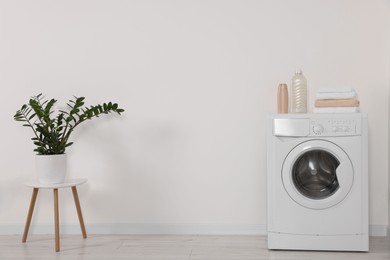 Photo of Modern washing machine and potted plant on stool against white wall indoors