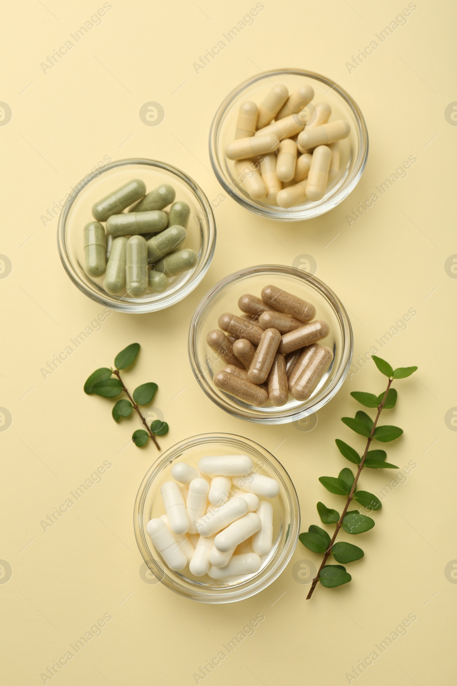 Photo of Different vitamin capsules in bowls and branches on pale yellow background, flat lay