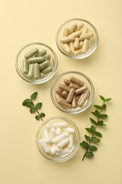 Different vitamin capsules in bowls and branches on pale yellow background, flat lay