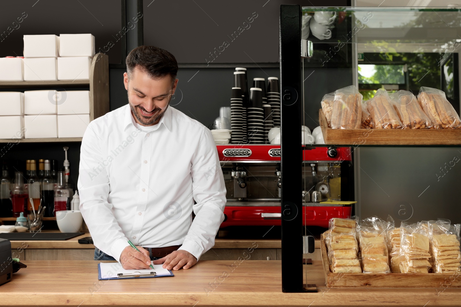 Photo of Business owner in his cafe. Man writing notes on clipboard near showcase with pastries