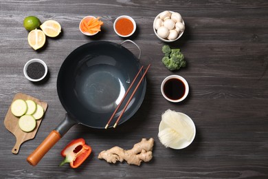 Photo of Empty iron wok and chopsticks surrounded by ingredients on dark grey wooden table, flat lay. Space for text