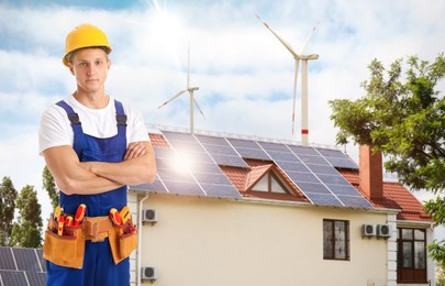 Image of Worker with tools wearing uniform and view of wind energy turbines near house with installed solar panels on roof