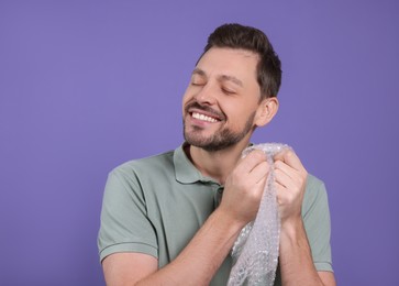 Photo of Happy man popping bubble wrap on purple background, space for text. Stress relief