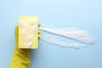 Cleaner in rubber glove holding sponge with foam on light blue background, top view