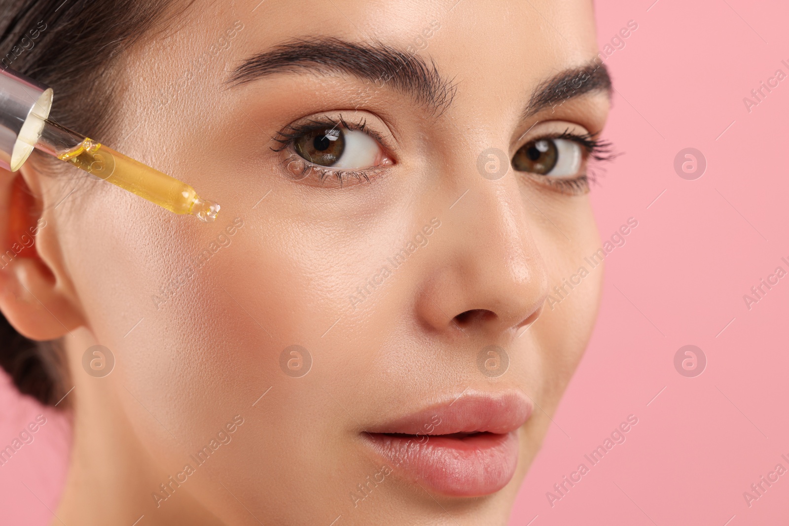 Photo of Beautiful young woman applying serum onto her face on pink background, closeup