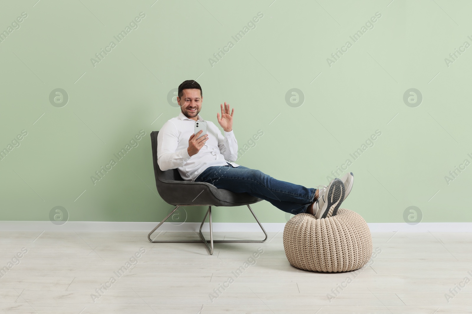 Photo of Happy man sitting in armchair and having video chat via smartphone near light green wall indoors