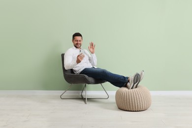 Happy man sitting in armchair and having video chat via smartphone near light green wall indoors