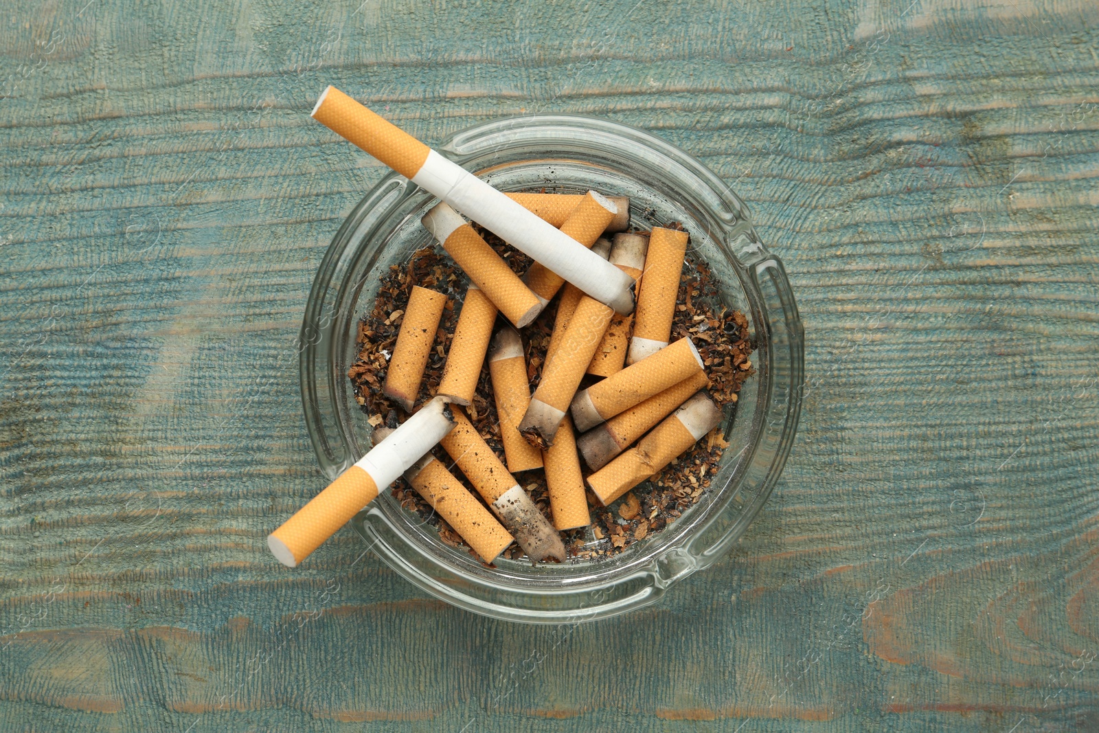 Photo of Glass ashtray with cigarette stubs on light blue wooden table, top view