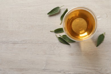 Glass cup of sage tea and green leaves on wooden table, top view. Space for text