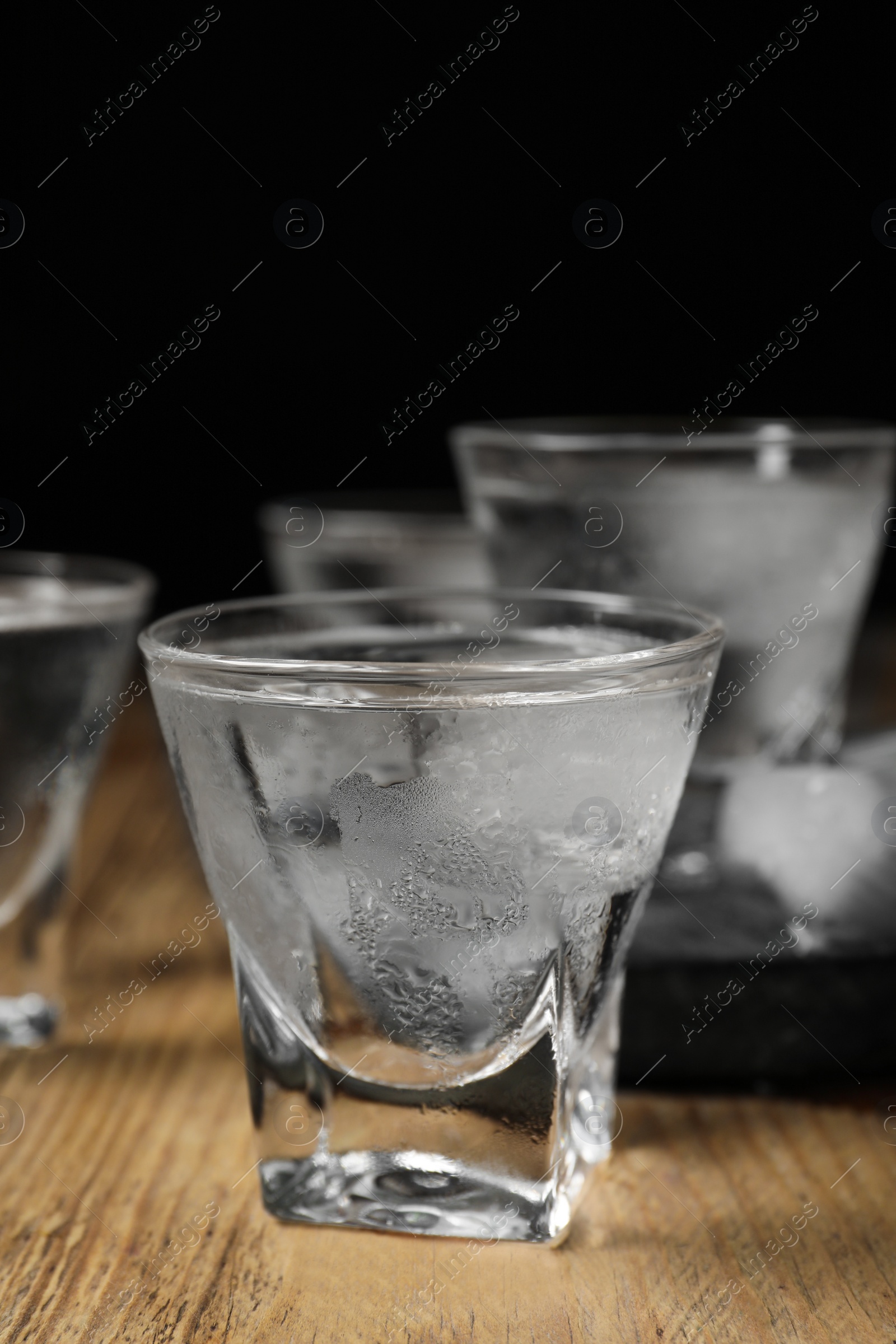 Photo of Shot glasses of vodka with ice cubes on wooden table against black background, closeup