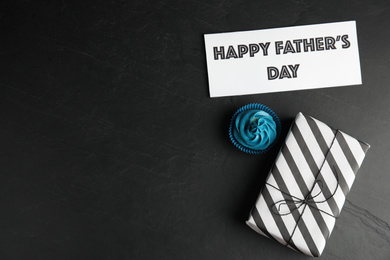 Photo of Cupcake, gift box and card with phrase HAPPY FATHER'S DAY on black table, flat lay. Space for text