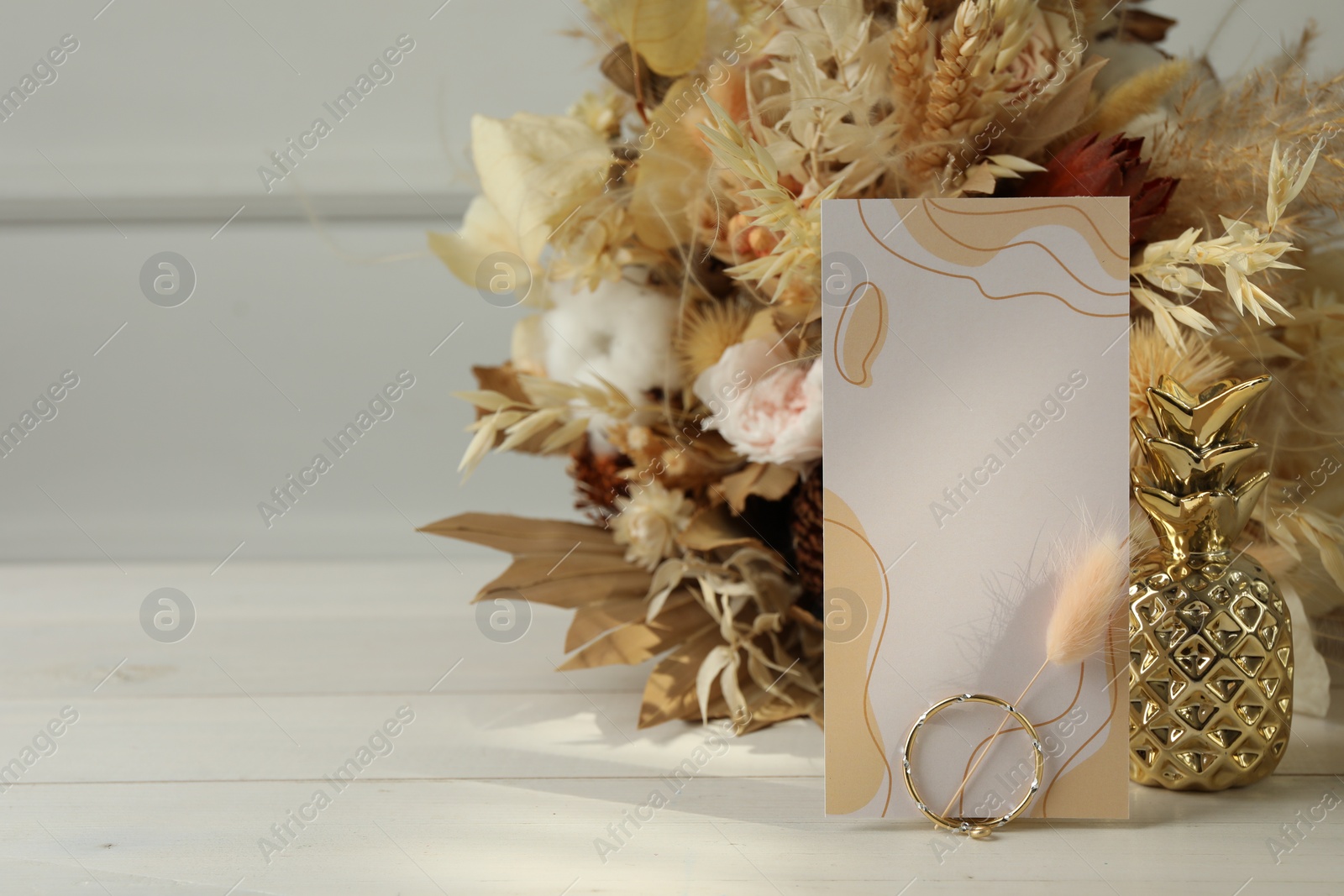 Photo of Blank invitation card, ring, dry leaves and flowers on white wooden table. Space for text