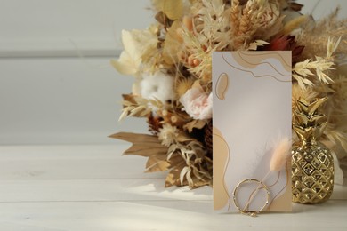 Blank invitation card, ring, dry leaves and flowers on white wooden table. Space for text