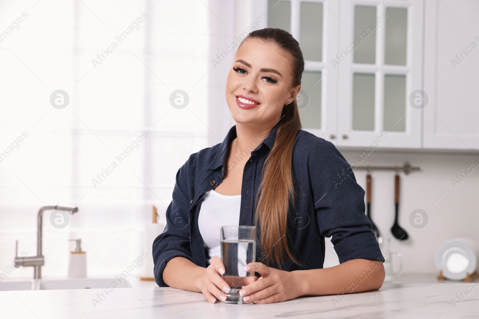 Photo of Happy woman with glass of fresh water at white marble table in kitchen