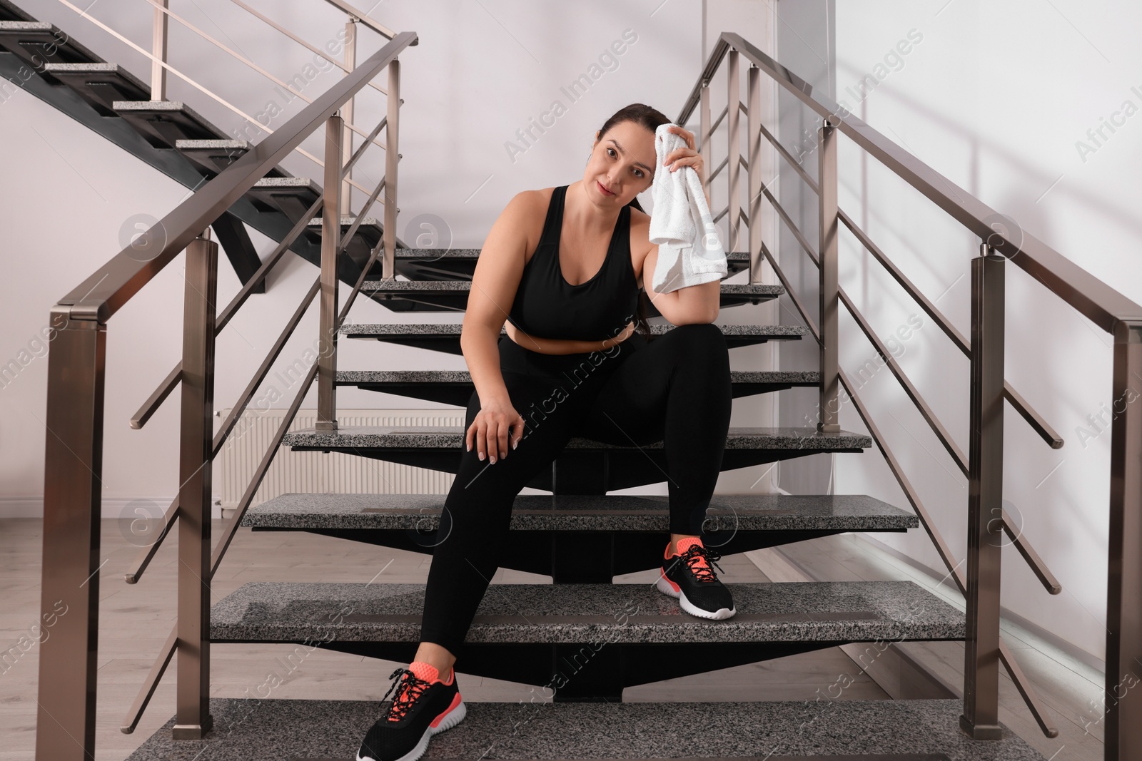 Photo of Tired overweight woman with towel sitting on stairs indoors