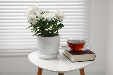 Photo of Beautiful chrysanthemum plant in flower pot, cup of tea and book on white table indoors