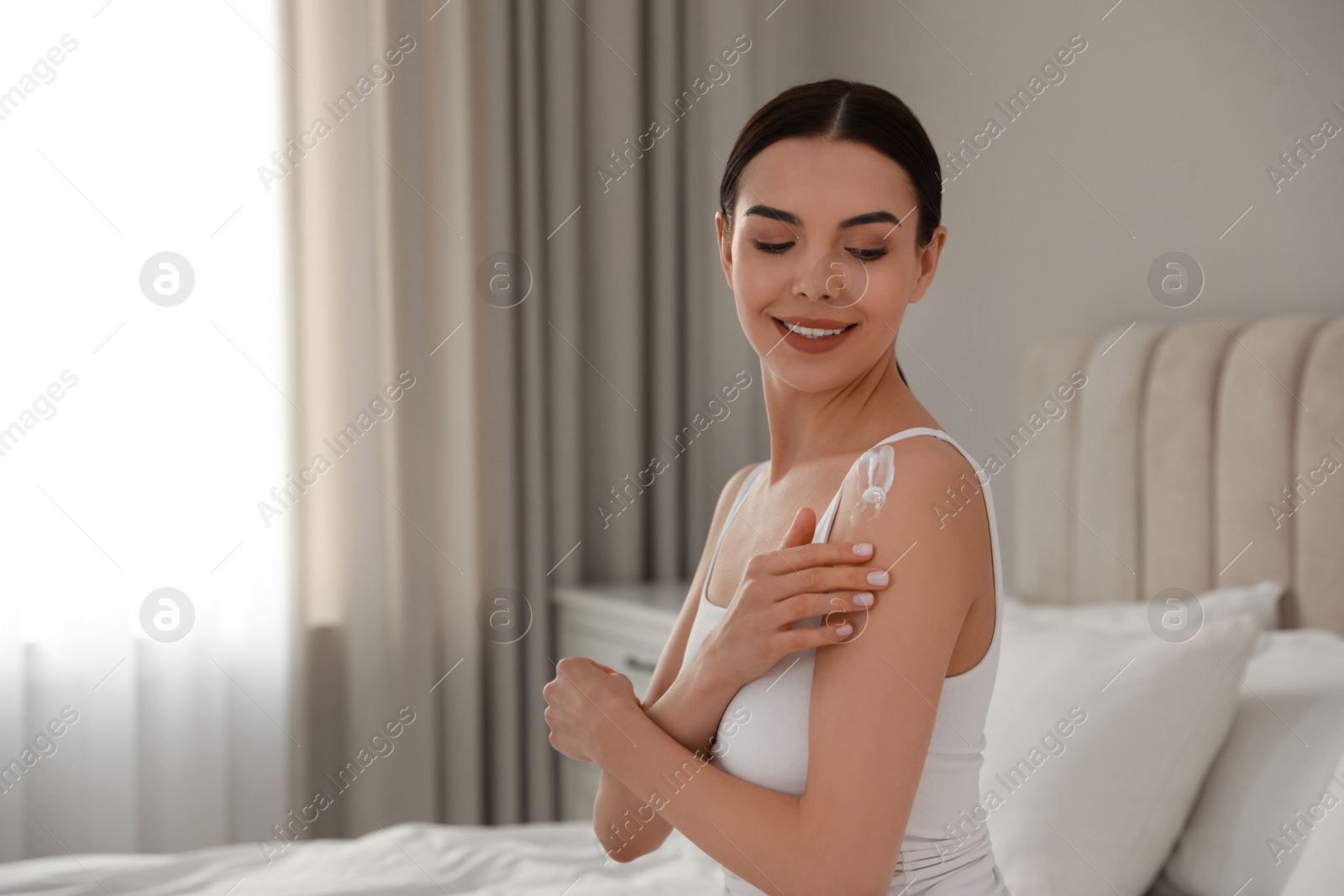 Photo of Young woman applying body cream onto her shoulder at home, space for text