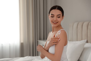 Photo of Young woman applying body cream onto her shoulder at home, space for text