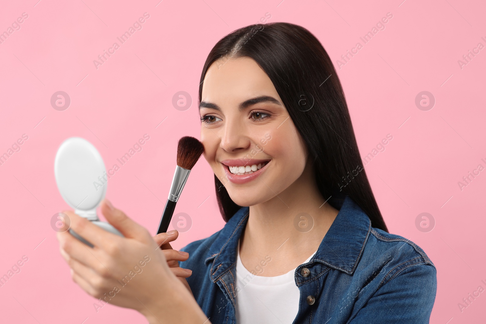 Photo of Beautiful woman with cosmetic pocket mirror applying makeup on pink background