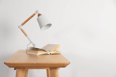 Photo of Stylish modern desk lamp and open book on wooden table near white wall, space for text