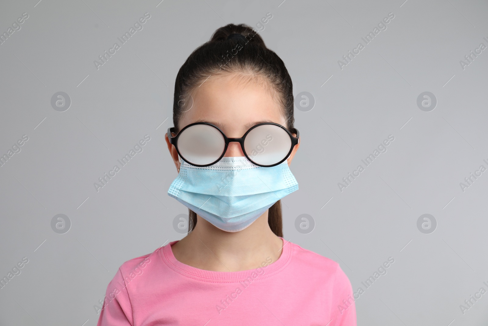 Photo of Little girl with foggy glasses caused by wearing medical face mask on grey background. Protective measure during coronavirus pandemic