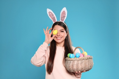 Beautiful woman in bunny ears headband holding basket with Easter eggs on color background