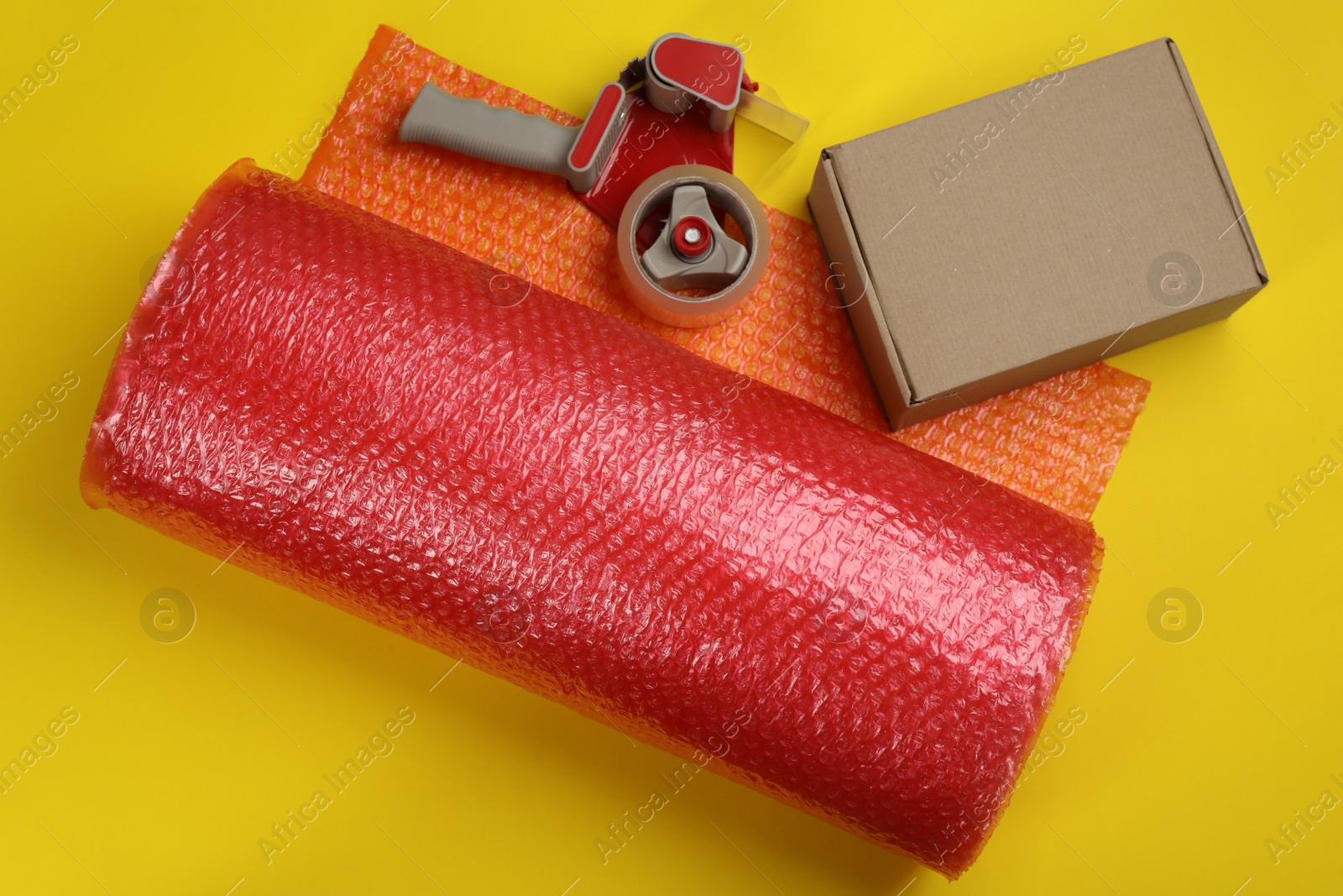 Photo of Bubble wrap roll, cardboard box and tape dispenser on yellow background, flat lay