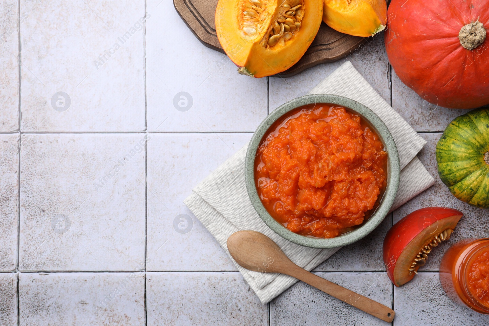 Photo of Bowl of delicious pumpkin jam and fresh pumpkins on tiled surface, flat lay. Space for text