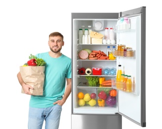 Young man with bag of groceries near open refrigerator on white background