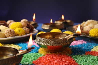 Diwali celebration. Diya lamps and colorful rangoli on table against violet background, closeup