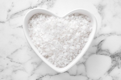 Photo of Heart shaped bowl with white sea salt on marble table, top view. Spa treatment