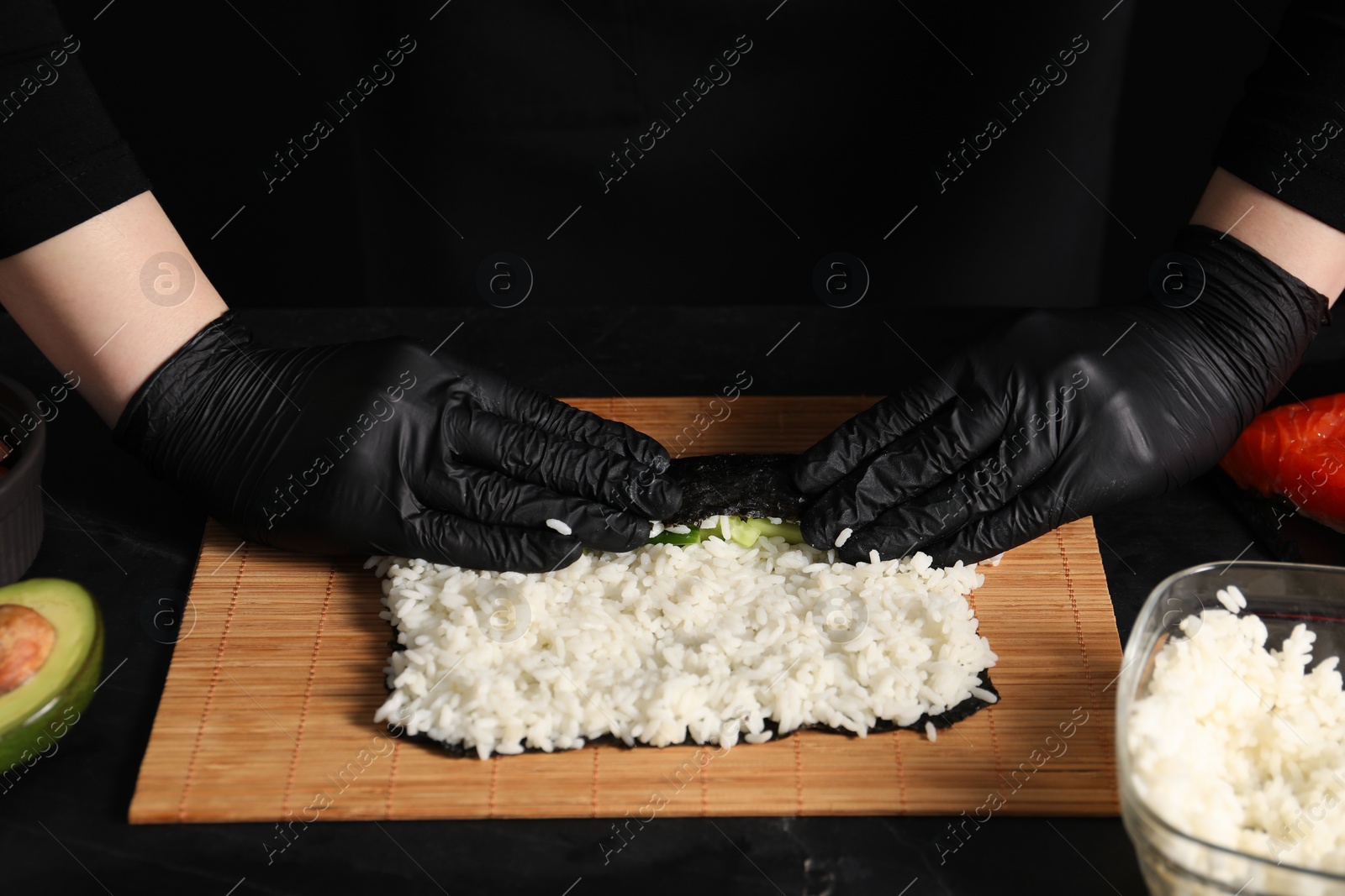 Photo of Chef in gloves wrapping sushi roll at dark table, closeup