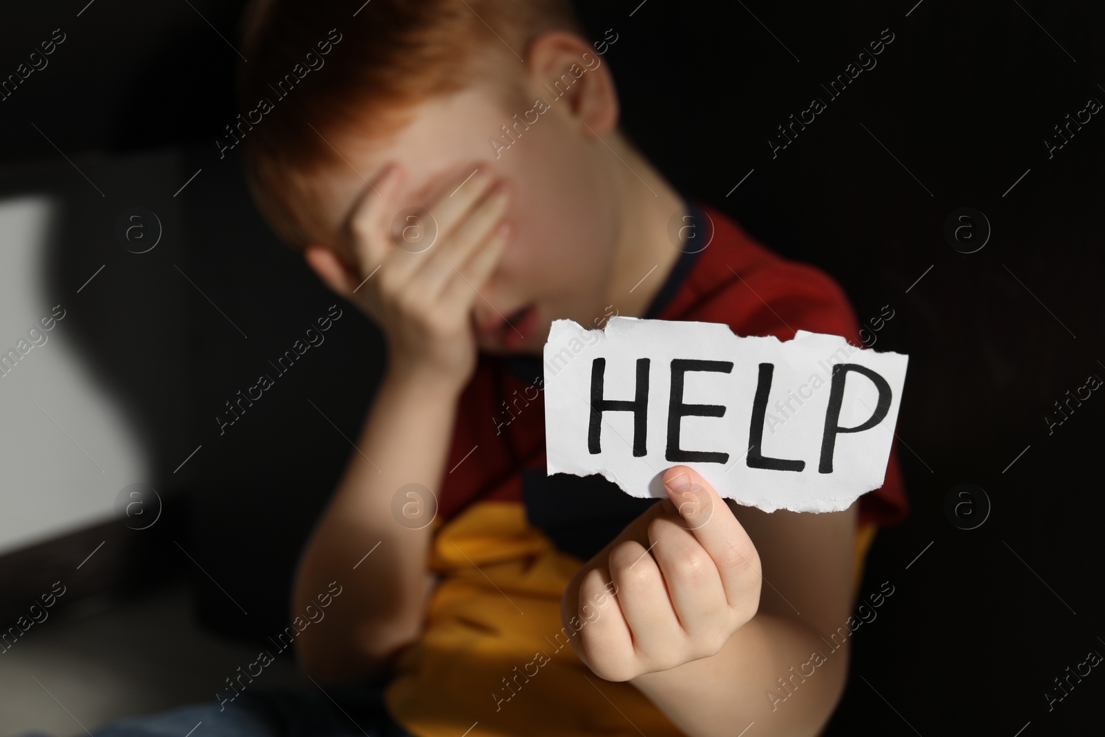 Photo of Little boy holding piece of paper with word Help under table, focus on hand. Domestic violence concept