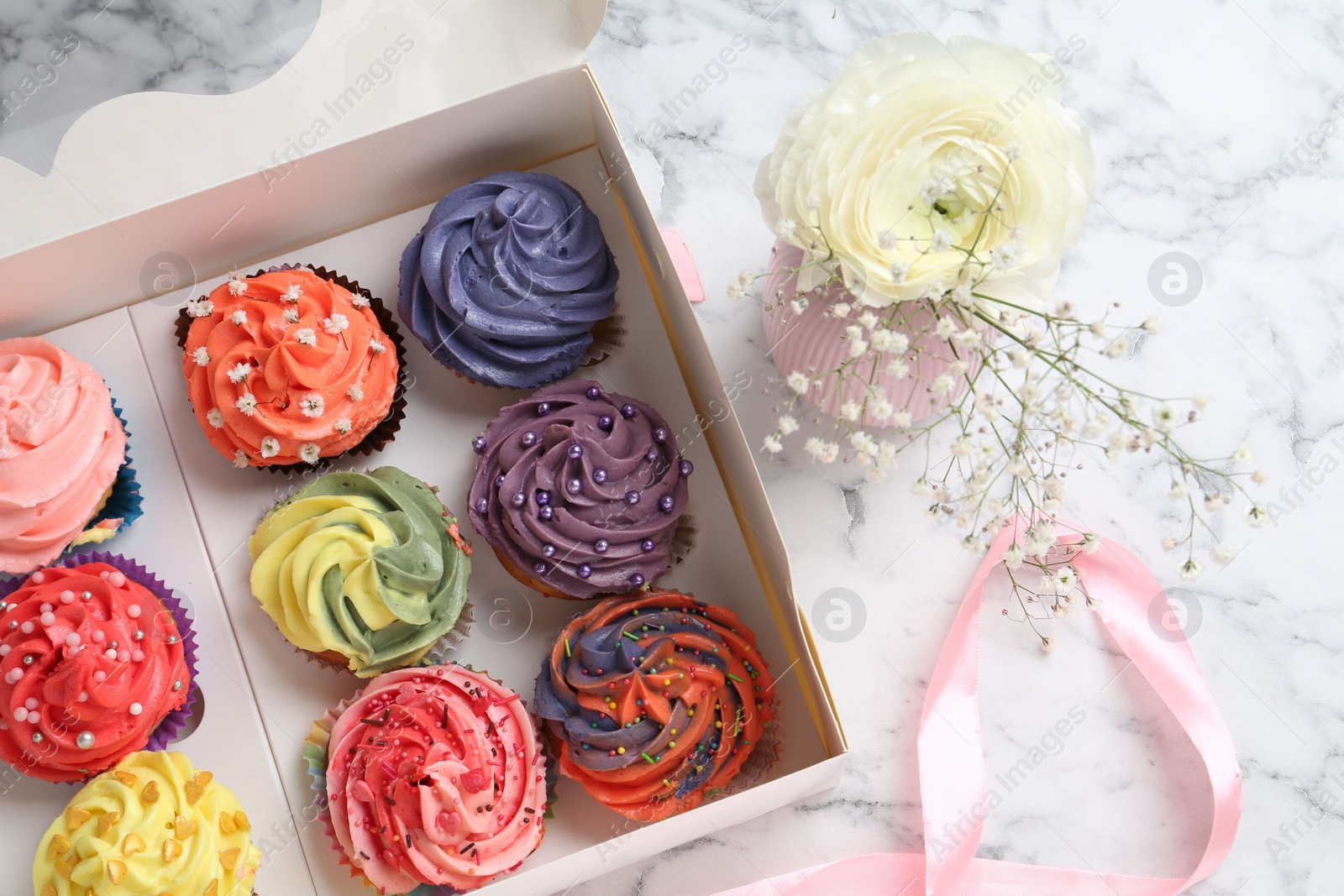 Photo of Different colorful cupcakes in box and flowers on white marble table, top view