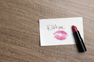 Photo of Paper card with lip print, phrase Kiss Me and lipstick on wooden table, top view. Space for text