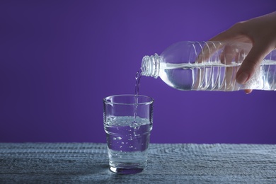 Photo of Woman pouring water from bottle into glass on blue wooden table against purple background, closeup