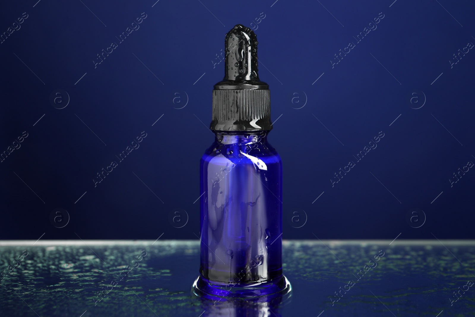 Photo of Bottle of face serum on wet surface against blue background, closeup