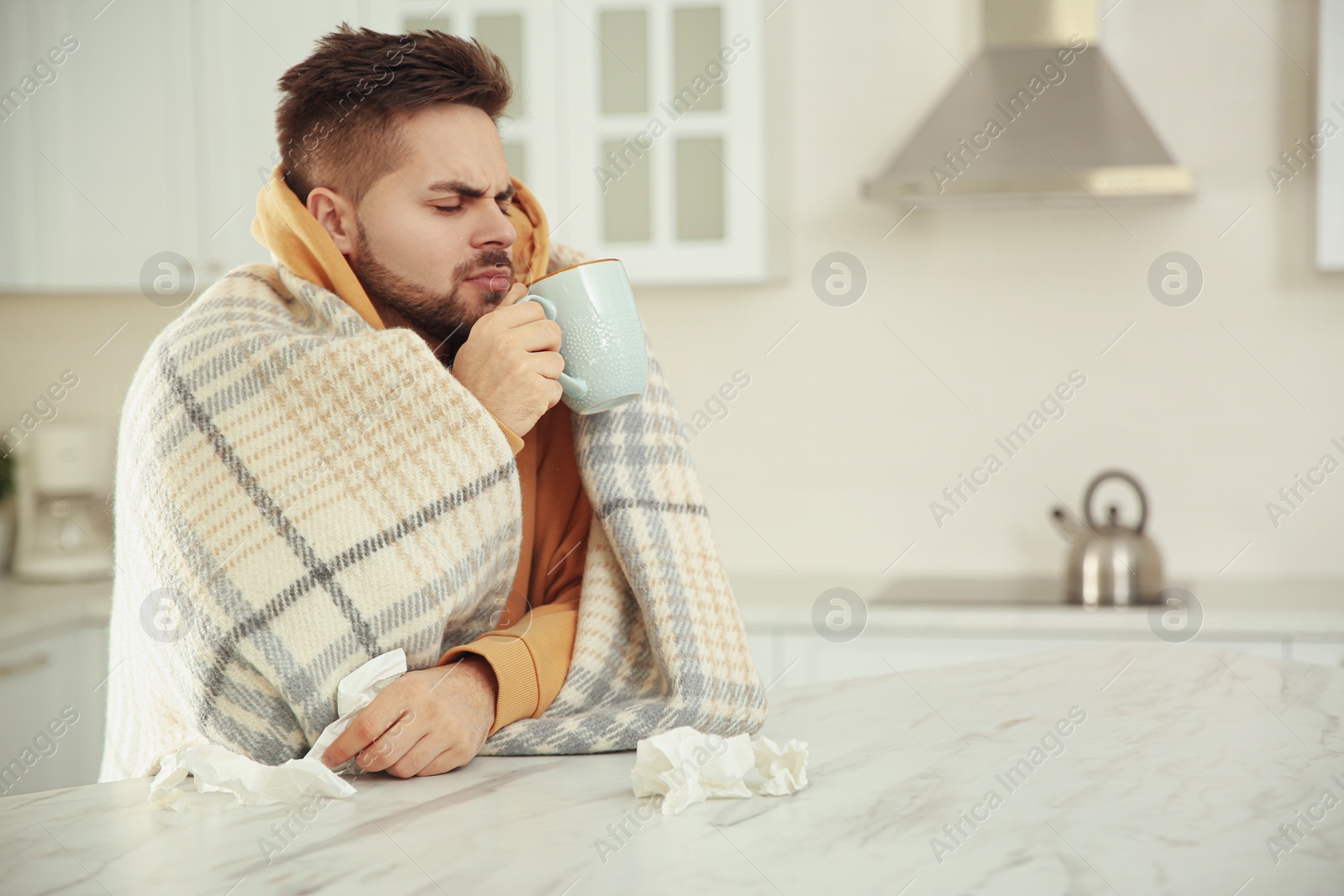 Photo of Sick young man with cup of hot drink and tissues in kitchen. Influenza virus